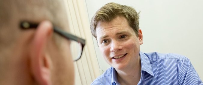 An image of two sitting men in conversation, focused on a fair-haired man wearing a light blue shirt. 
